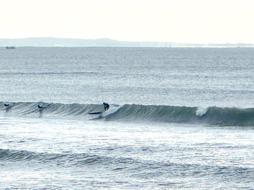 10月23日10時前の湘南茅ヶ崎の海。横に長く続く波の上で1人がボードに立って身をかがめ、左の海面にも人の姿があり、奥には陸地がかすんで見える